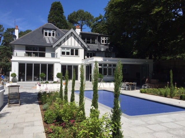 Large white house with multiple gables and large windows, set in a garden with a rectangular pool and manicured plants.