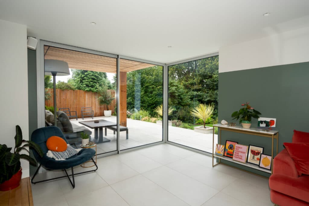 Modern living room with large corner windows overlooking a garden. Interior features a blue chair, red sofa, potted plants, and framed artwork on a console table. Patio view with outdoor seating.