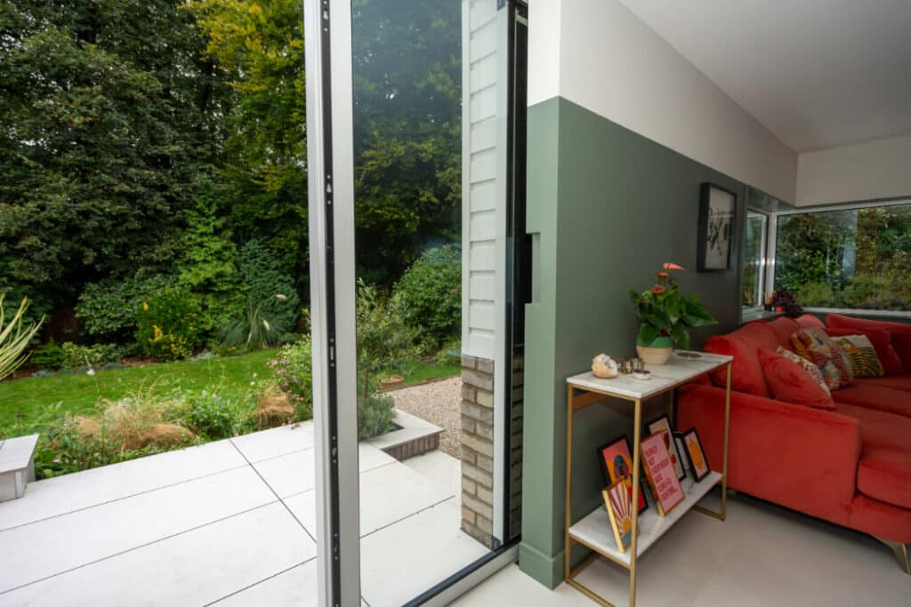 Room interior with a red sofa and a small table with decorations. Large glass doors open to a green garden with a paved patio area.