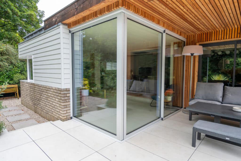 Modern outdoor patio with large glass sliding doors, wood-paneled overhang, and cushioned seating. Brick and wood exterior with a view of a garden pathway.