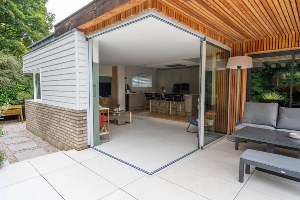 Modern home with open sliding doors showing a bright living area, kitchen with bar stools, and outdoor seating on a patio.