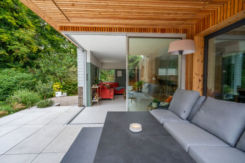 Modern outdoor patio with a gray couch and table, leading through sliding glass doors to an indoor living area with a red sofa. Greenery visible outside.