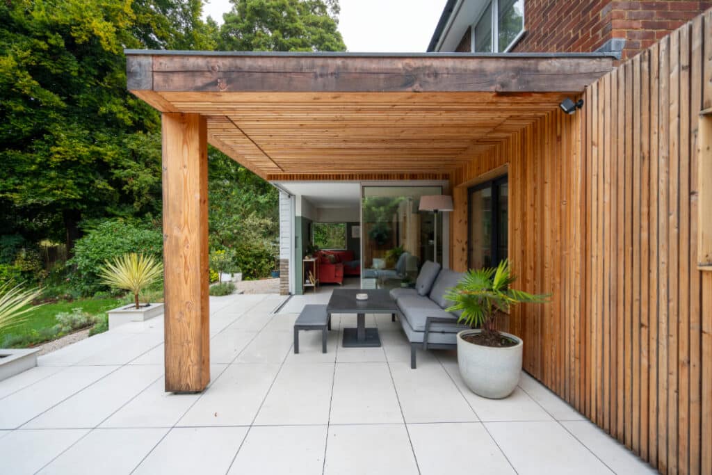 Wooden outdoor patio with seating area, modern design. Surrounded by potted plants and trees, leading into a house with glass doors.