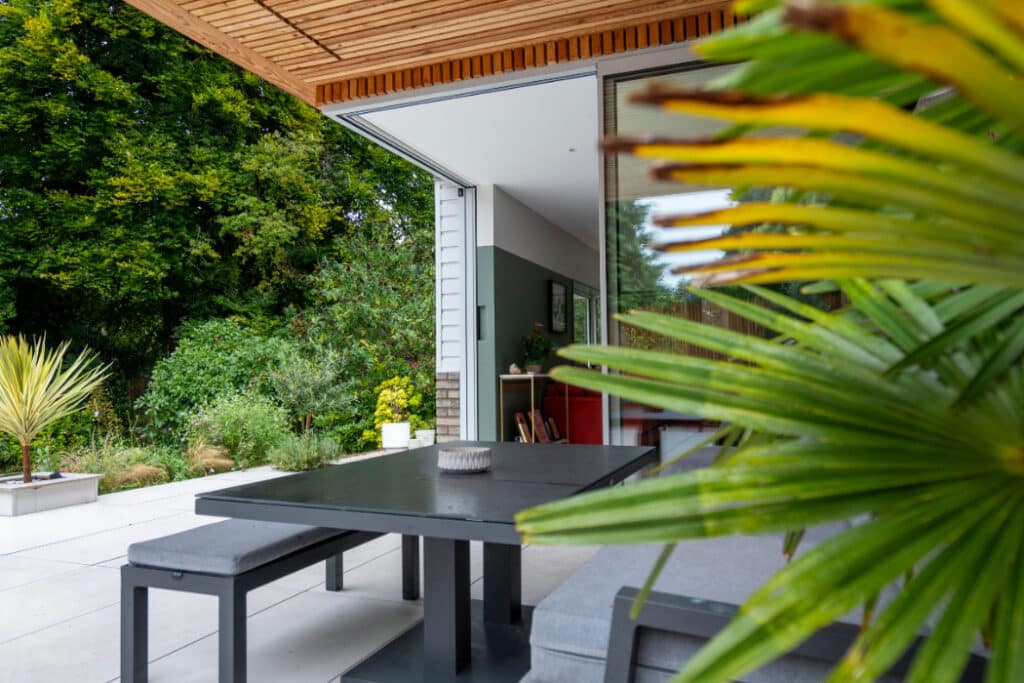 Modern outdoor patio with a black table and benches under a wooden overhang, surrounded by greenery and large plants. Open sliding glass door leads to the interior.