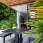 Thumbnail of http://Modern%20outdoor%20patio%20with%20a%20black%20table%20and%20benches%20under%20a%20wooden%20overhang,%20surrounded%20by%20greenery%20and%20large%20plants.%20Open%20sliding%20glass%20door%20leads%20to%20the%20interior.