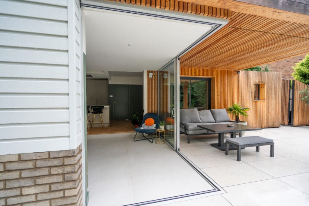 Modern patio with open sliding glass doors revealing indoor and outdoor seating areas. Features a sofa, table, and plants on a tiled floor. Walls include wooden paneling and white siding.