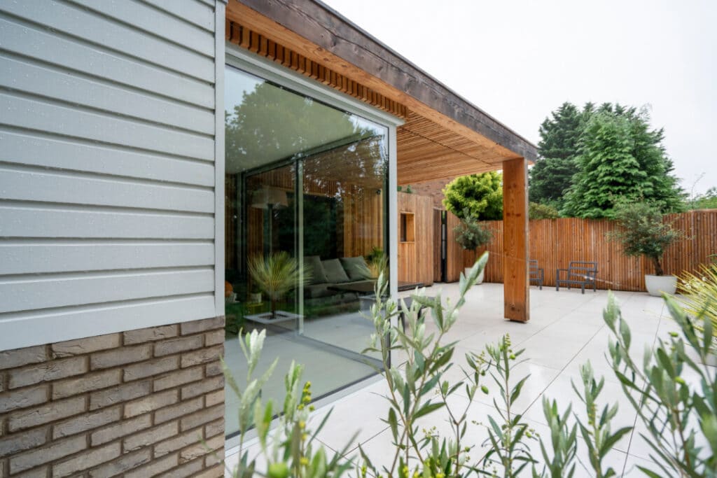 Modern patio with glass sliding doors, wooden overhang, and white brick exterior. Outdoor seating and greenery visible, with trees in the background.