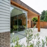 Thumbnail of http://Modern%20patio%20with%20glass%20sliding%20doors,%20wooden%20overhang,%20and%20white%20brick%20exterior.%20Outdoor%20seating%20and%20greenery%20visible,%20with%20trees%20in%20the%20background.