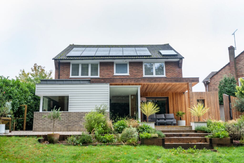 Two-story brick house with a modern extension, solar panels on the roof, a wooden patio, and a landscaped garden with various plants.