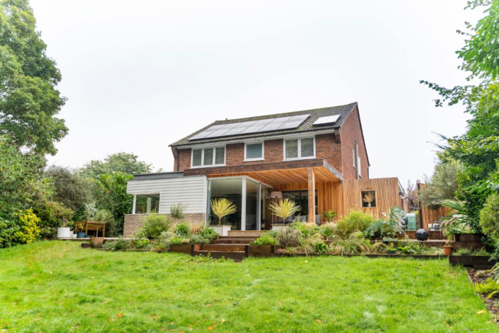 A two-story brick house with solar panels and a wooden extension, surrounded by a garden with green grass and various plants.