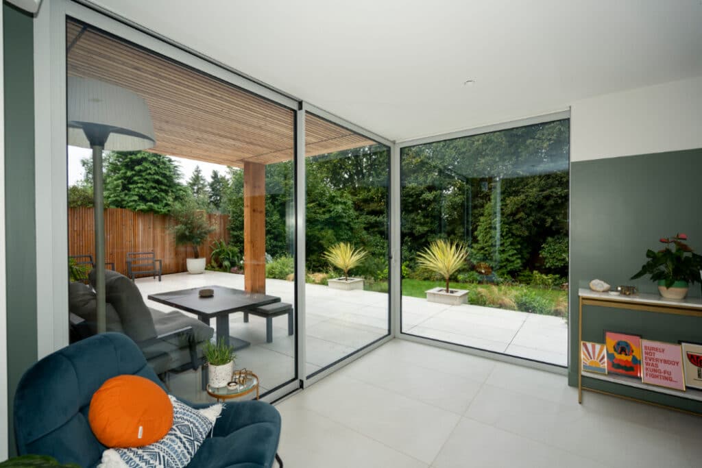 Modern interior with a large glass door opening to a patio and garden. A blue armchair with an orange cushion is in the foreground, and a lamp and side table are nearby.