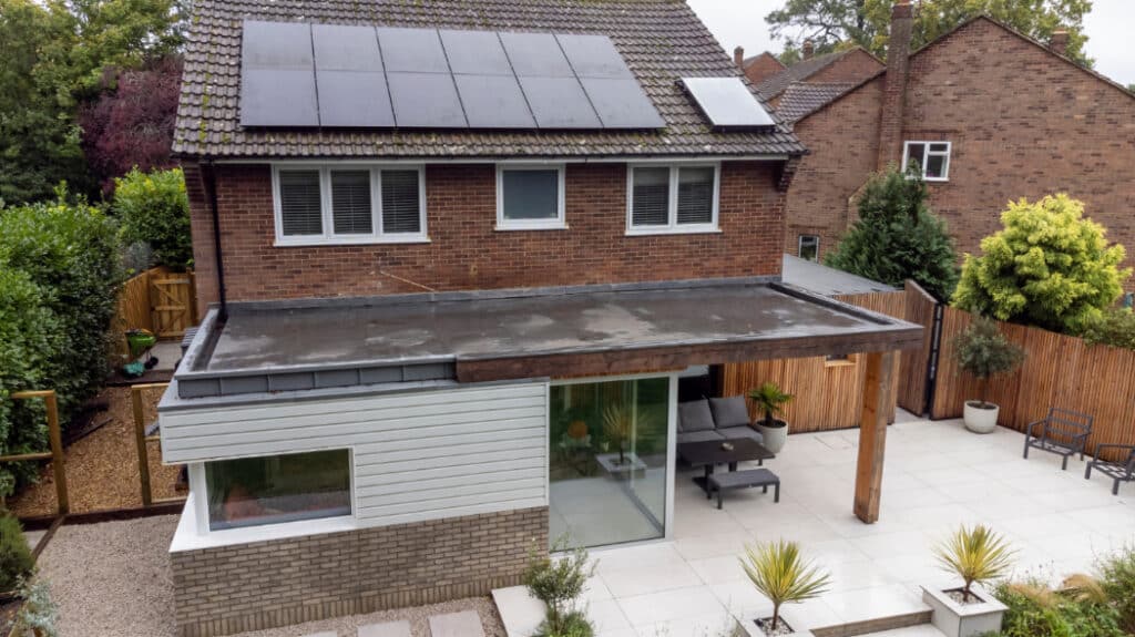 A modern two-story house with solar panels on the roof and a flat-roof extension featuring large windows. The backyard has a paved patio area with outdoor furniture.