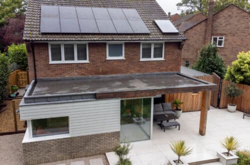 A modern two-story house with solar panels on the roof and a flat-roof extension featuring large windows. The backyard has a paved patio area with outdoor furniture.