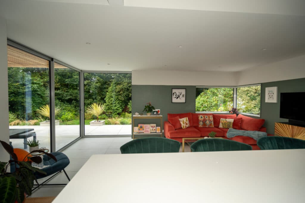 Modern living room with large glass doors, red sectional sofa, green accent wall, and visible greenery outside.