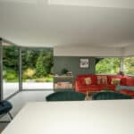 Thumbnail of http://Modern%20living%20room%20with%20large%20glass%20doors,%20red%20sectional%20sofa,%20green%20accent%20wall,%20and%20visible%20greenery%20outside.