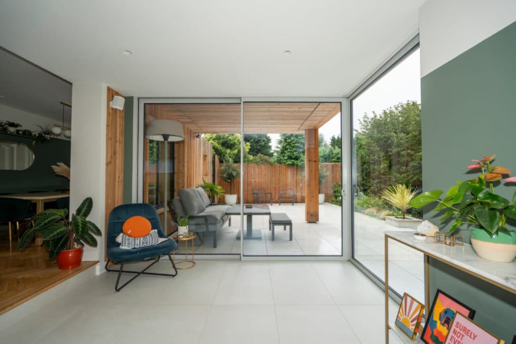 Modern living room with glass sliding doors leading to a patio. Indoor plants on the left and right, furniture outside, and greenery in the background. Tile flooring throughout.