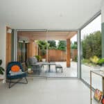 Thumbnail of http://Modern%20living%20room%20with%20glass%20sliding%20doors%20leading%20to%20a%20patio.%20Indoor%20plants%20on%20the%20left%20and%20right,%20furniture%20outside,%20and%20greenery%20in%20the%20background.%20Tile%20flooring%20throughout.