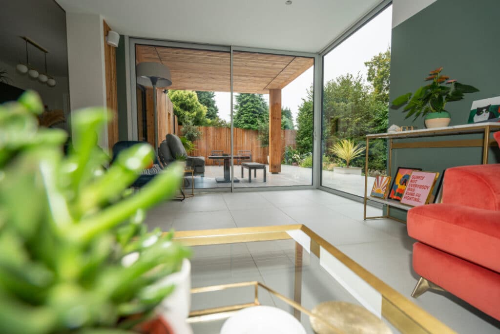 Modern living room with large glass doors opening to a garden. Features a red chair, green plant, and abstract artwork. Outdoor area has seating and wooden fencing.