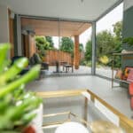 Thumbnail of http://Modern%20living%20room%20with%20large%20glass%20doors%20opening%20to%20a%20garden.%20Features%20a%20red%20chair,%20green%20plant,%20and%20abstract%20artwork.%20Outdoor%20area%20has%20seating%20and%20wooden%20fencing.