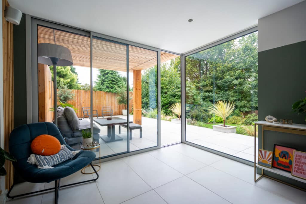 Spacious room with large glass doors leading to a patio. Modern furniture includes a blue chair and a side table with decor. Outdoor patio has a table, chairs, and lush greenery.