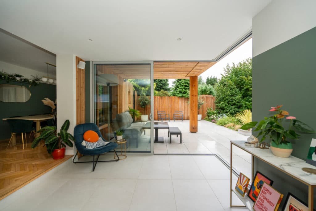 Modern indoor space with large glass doors opening to a patio. Features a chair, potted plant, and a view of a garden with trees and a wooden fence.