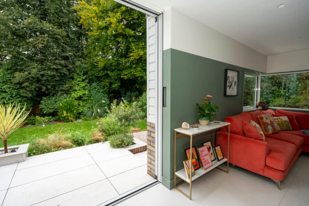 A modern living room with a red sofa and decorative items on a shelf. Sliding doors open to a garden with green foliage and a paved patio area.