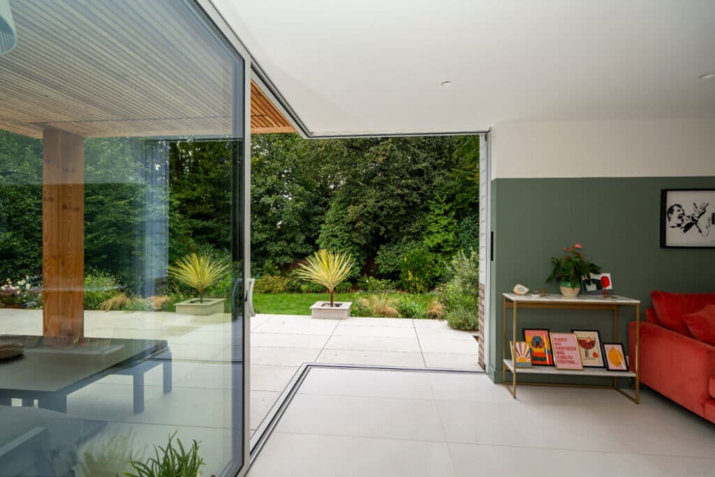 Modern living room with large glass sliding doors leading to a patio and lush garden. Red couch and small shelf with plants and framed pictures inside. Minimalist and airy design.