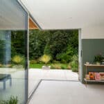 Thumbnail of http://Modern%20living%20room%20with%20large%20glass%20sliding%20doors%20leading%20to%20a%20patio%20and%20lush%20garden.%20Red%20couch%20and%20small%20shelf%20with%20plants%20and%20framed%20pictures%20inside.%20Minimalist%20and%20airy%20design.