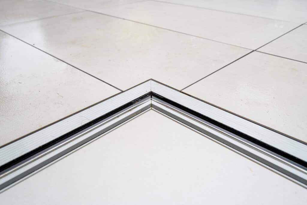 Close-up of a tiled floor with a metal strip at the junction of two corners. The tiles are light-colored and have a glossy finish.