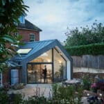 Thumbnail of http://Modern%20glass%20extension%20with%20metal%20roof%20on%20a%20traditional%20brick%20house,%20surrounded%20by%20a%20garden%20with%20various%20plants%20and%20flowers;%20a%20person%20is%20visible%20inside%20through%20the%20large%20windows.