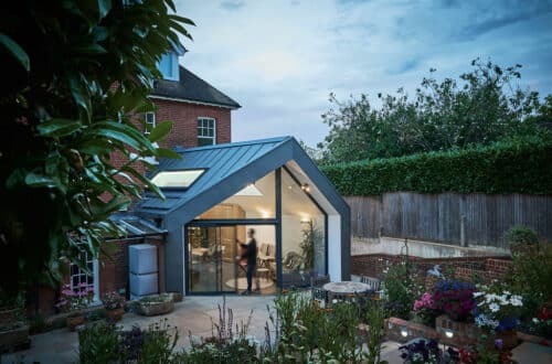 Modern glass extension with metal roof on a traditional brick house, surrounded by a garden with various plants and flowers; a person is visible inside through the large windows.