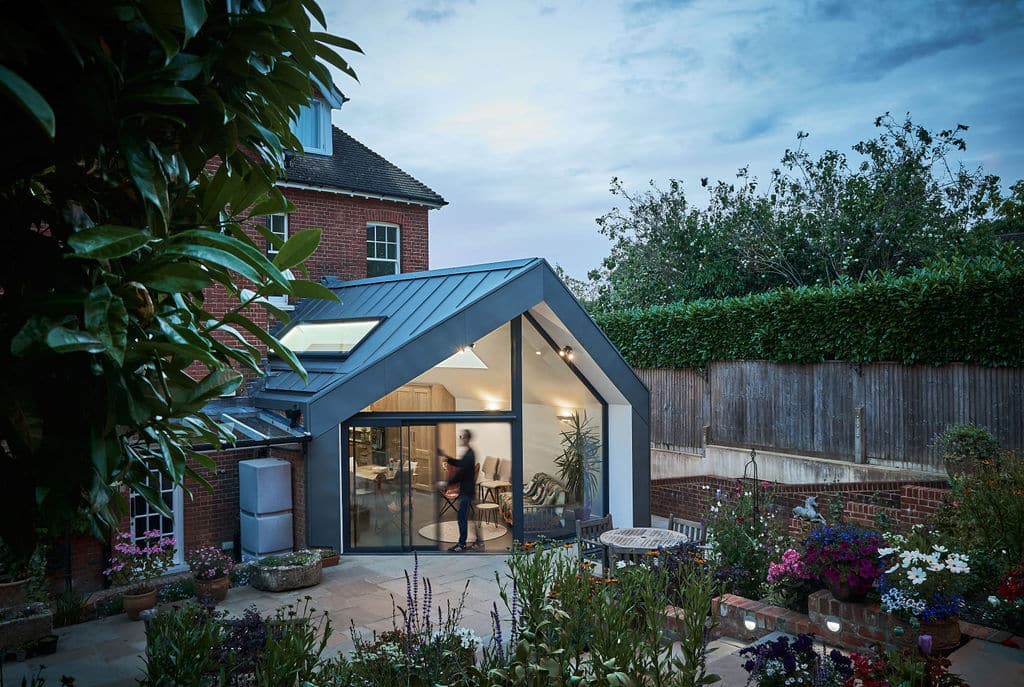 Modern glass extension with metal roof on a traditional brick house, surrounded by a garden with various plants and flowers; a person is visible inside through the large windows.