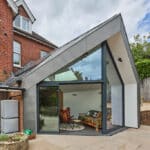 Thumbnail of http://Modern%20glass%20extension%20with%20angular%20roof%20attached%20to%20a%20red%20brick%20house.%20Interior%20features%20colorful%20seating%20and%20a%20potted%20plant.%20Outside,%20there%20is%20a%20wooden%20fence%20and%20a%20small%20patio.