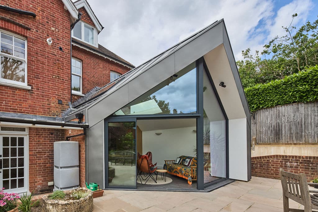 Modern glass extension with angular roof attached to a red brick house. Interior features colorful seating and a potted plant. Outside, there is a wooden fence and a small patio.