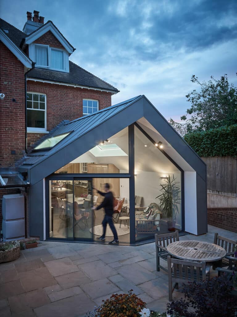 A modern house extension with large glass doors and a high ceiling. A person is seen walking inside. There is outdoor seating on the patio in the foreground.