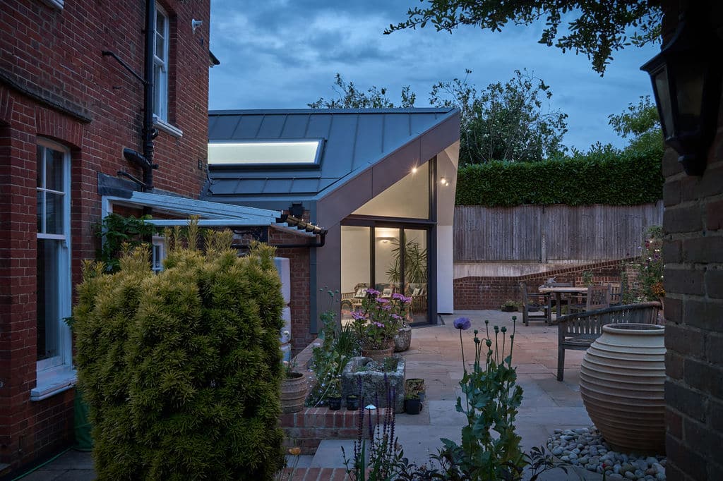 Modern extension with large glass sliding doors attached to a brick building, surrounded by a garden patio with plants and outdoor furniture, under a twilight sky.