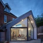 Thumbnail of http://Modern%20house%20extension%20with%20large%20glass%20windows%20and%20an%20angular%20roof%20design,%20showing%20a%20well-lit%20interior%20with%20furniture%20and%20plants,%20adjacent%20to%20an%20older%20brick%20building.