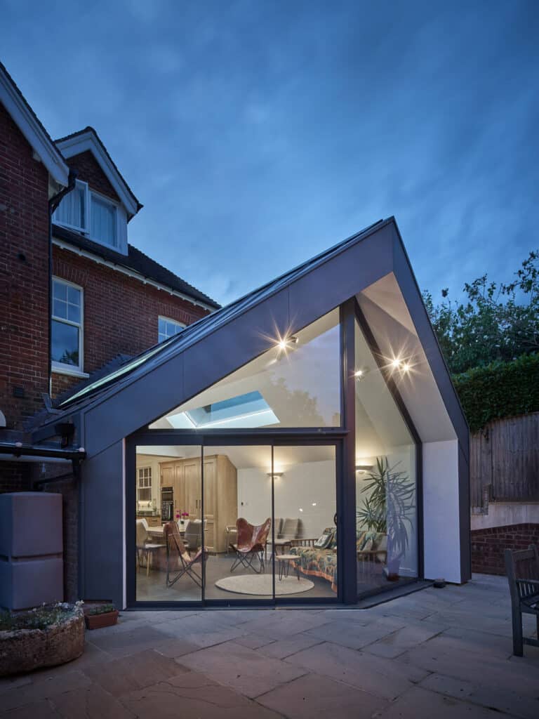 Modern house extension with large glass windows and an angular roof design, showing a well-lit interior with furniture and plants, adjacent to an older brick building.