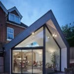 Thumbnail of http://Modern%20extension%20with%20large%20glass%20windows%20and%20angular%20roof%20attached%20to%20a%20traditional%20brick%20house,%20interior%20visible%20with%20plants%20and%20seating,%20patio%20in%20the%20foreground.