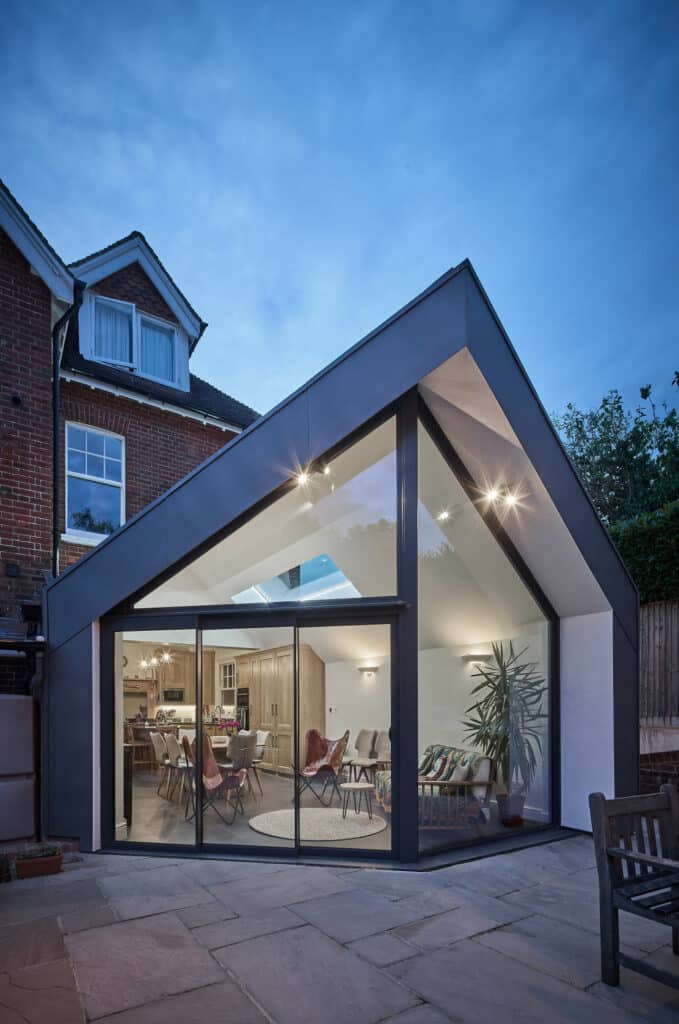 Modern extension with large glass windows and angular roof attached to a traditional brick house, interior visible with plants and seating, patio in the foreground.