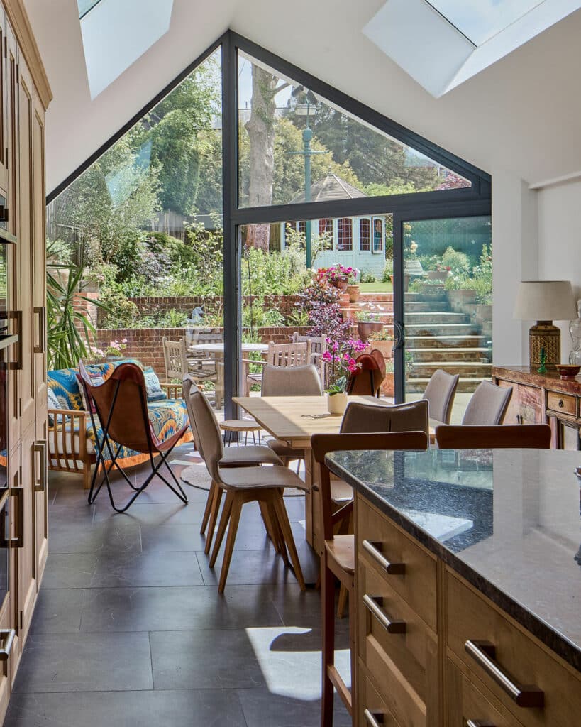 Stylish kitchen with large windows and skylights, modern wooden furniture, and a view of a lush garden with outdoor seating and steps leading to a house.