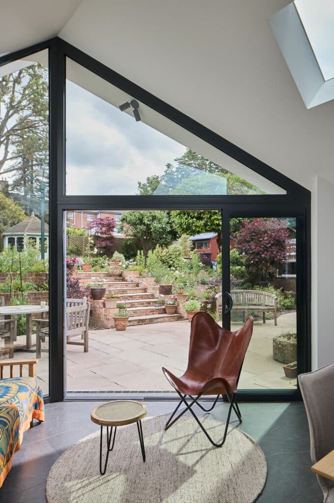 Room with a large triangular window overlooking a garden with steps and potted plants. Inside, there's a leather butterfly chair, small round table, and a rug on a wood floor.