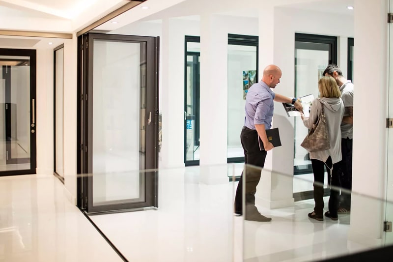 Three people stand in a modern, brightly lit showroom with large glass doors—possibly bifold doors—discussing or reviewing documents together in Scotland.