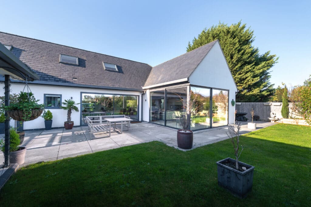 Modern house with large glass sliding doors opening to a patio with outdoor seating, surrounded by a well-kept lawn and potted plants, under a clear sky.