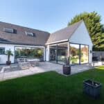 Thumbnail of http://Modern%20house%20with%20large%20glass%20sliding%20doors%20opening%20to%20a%20patio%20with%20outdoor%20seating,%20surrounded%20by%20a%20well-kept%20lawn%20and%20potted%20plants,%20under%20a%20clear%20sky.