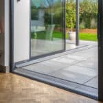 Thumbnail of http://A%20sliding%20glass%20door%20opens%20onto%20a%20tiled%20patio%20with%20outdoor%20furniture%20and%20a%20green%20lawn,%20viewed%20from%20a%20room%20with%20parquet%20flooring%20and%20a%20side%20table%20with%20a%20lamp.