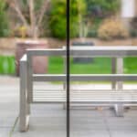 Thumbnail of http://A%20glass%20door%20divides%20the%20view%20of%20an%20outdoor%20patio%20with%20a%20white%20metal%20bench%20and%20table,%20with%20a%20green%20lawn%20and%20garden%20in%20the%20background.