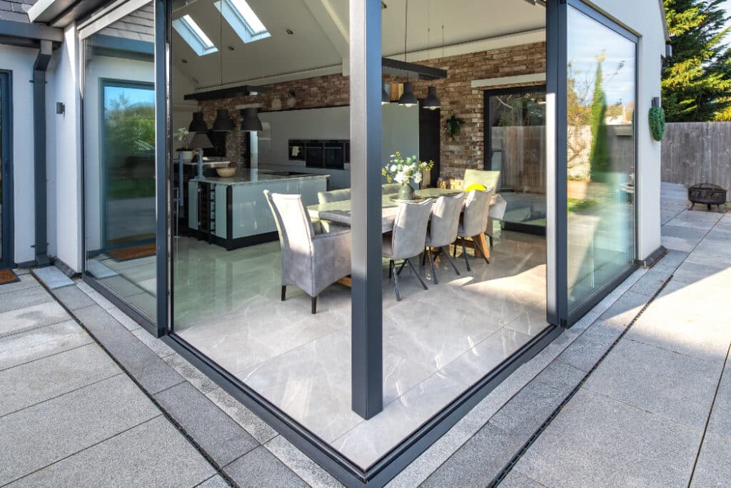 Modern dining room and kitchen with large glass walls, light grey tiled flooring, and contemporary furniture, viewed from outside on a tiled patio.
