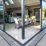 Thumbnail of http://Modern%20dining%20room%20and%20kitchen%20with%20large%20glass%20walls,%20light%20grey%20tiled%20flooring,%20and%20contemporary%20furniture,%20viewed%20from%20outside%20on%20a%20tiled%20patio.