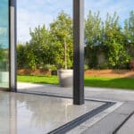 Thumbnail of http://View%20from%20a%20modern%20glass%20door%20opening%20to%20a%20patio%20with%20tiled%20flooring,%20green%20lawn,%20potted%20plant,%20and%20wooden%20raised%20garden%20bed%20with%20shrubs%20against%20a%20fence.